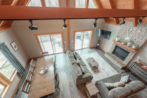 Un salón amplio con un sofá grande, una mesa de centro y una chimenea. Hay varias ventanas y un televisor montado en la pared.