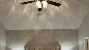 Un dormitorio con una cama grande, dos mesitas de noche con lámparas, un ventilador de techo y un cabecero capitonado.
