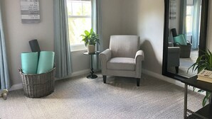 Una habitación con un sillón gris, una mesita auxiliar con una planta en maceta, una cesta de mimbre con objetos doblados y un espejo grande en la pared.