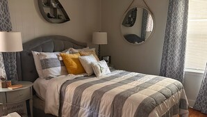 Una cama bien tendida con colcha a rayas, una mesita de noche con lámpara, un espejo montado en la pared y un ventilador de techo.
