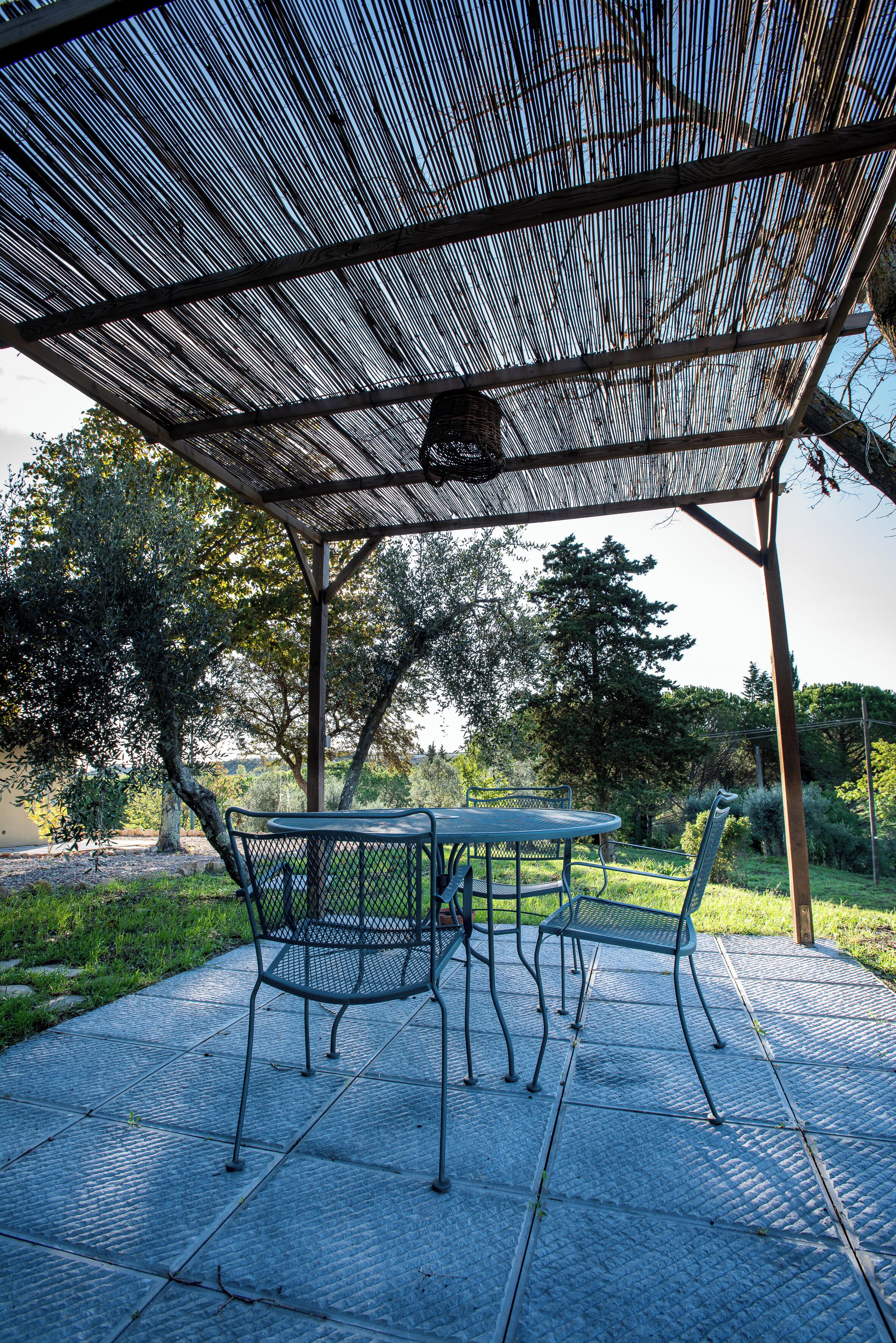 A covered outdoor seating area with a table and chairs, surrounded by trees and a clear sky.