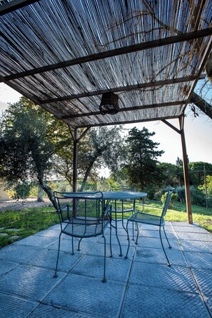A covered outdoor seating area with a table and chairs, surrounded by trees and a clear sky.