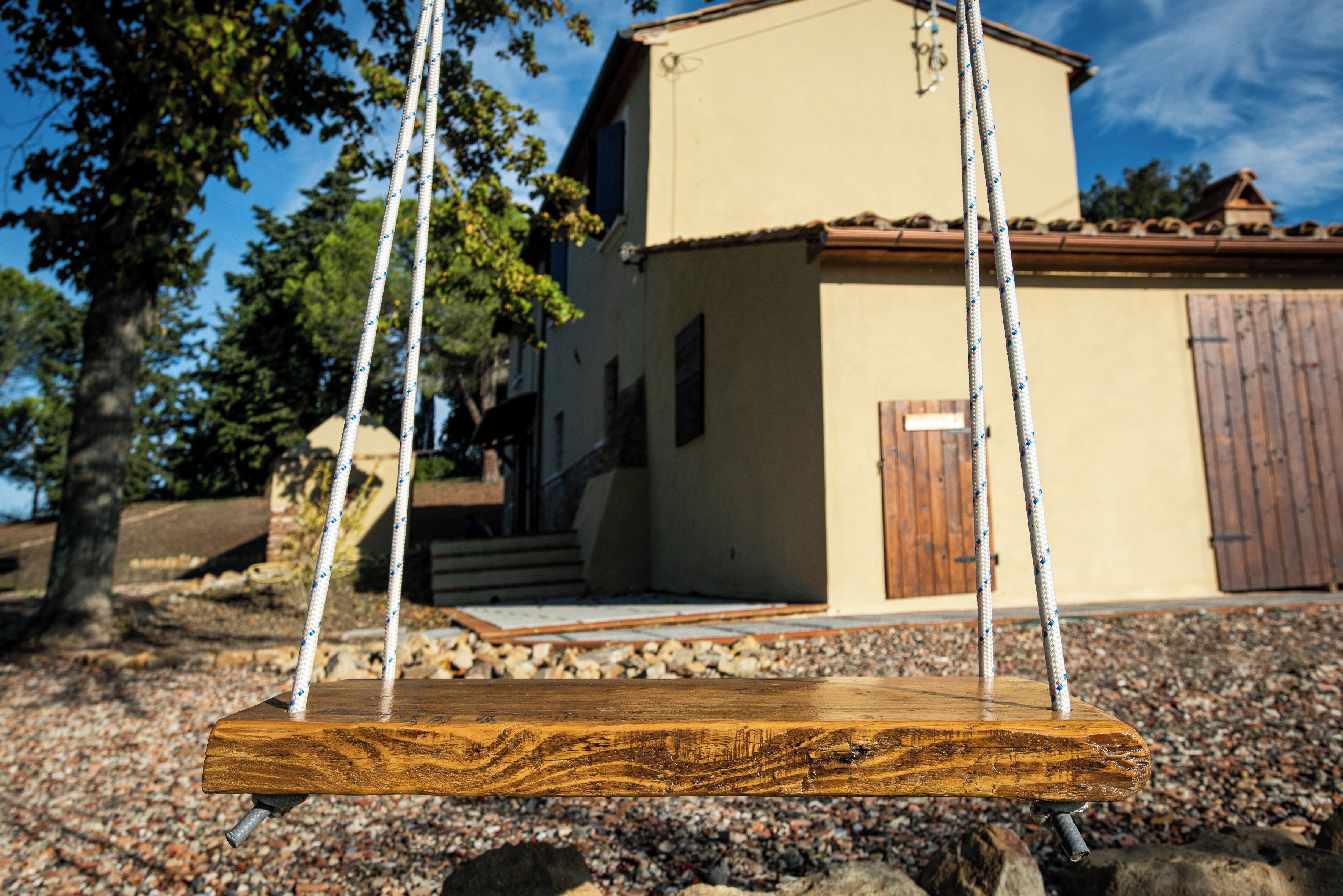 A wooden swing with ropes, a beige building with a brown door, and a gravel ground.