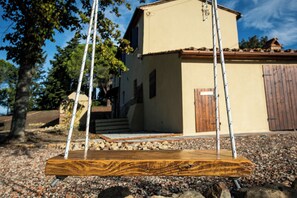Une balançoire en bois avec des cordes, un bâtiment de couleur beige avec une porte marron, et un sol recouvert de gravier.