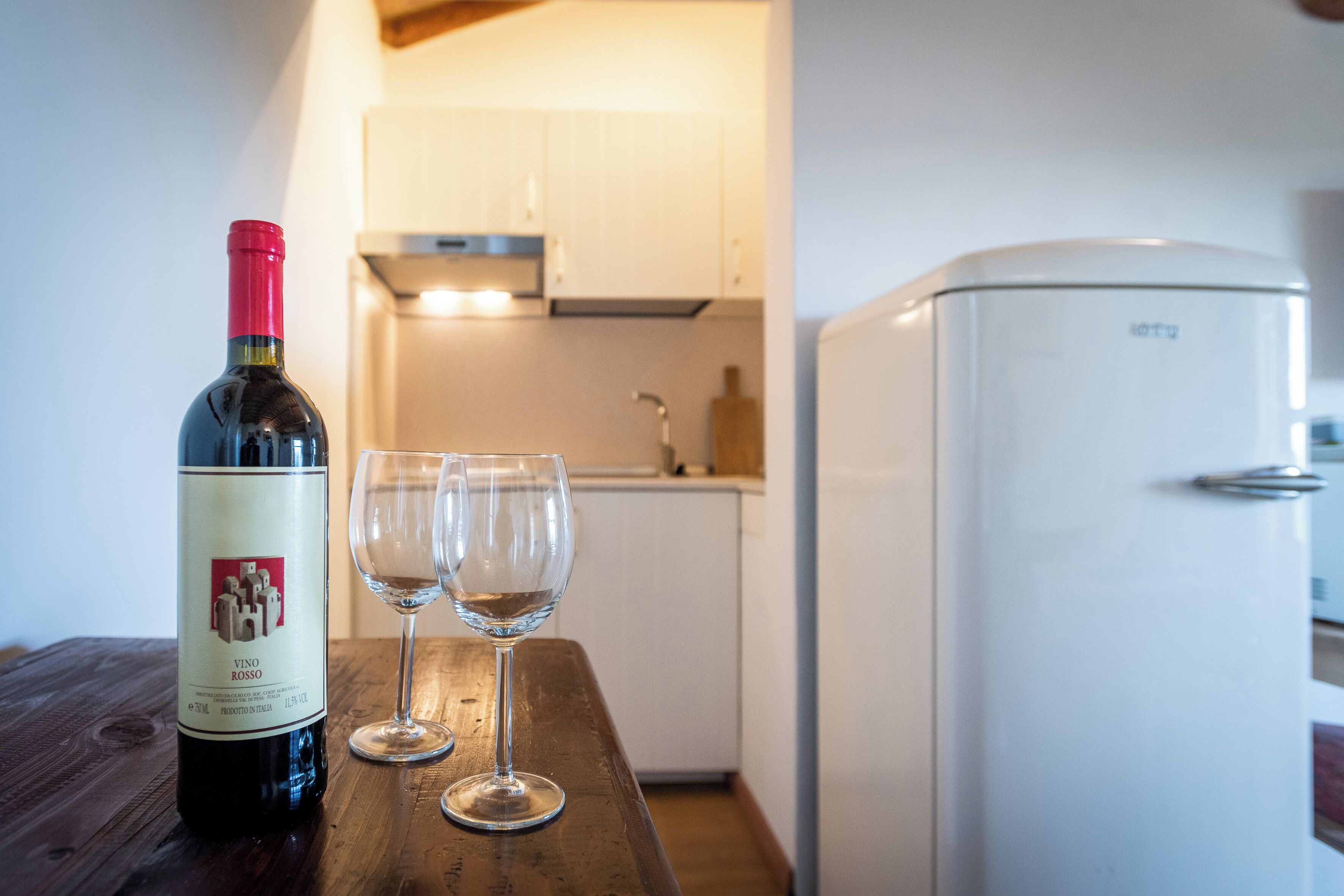 A bottle of wine and two glasses on a wooden table in a kitchen with white cabinets and a white refrigerator.