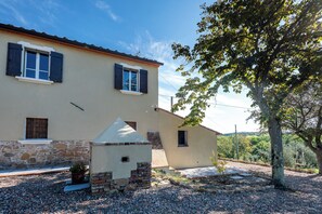Uma casa de dois andares com alicerce de pedra, uma construção pequena anexa e uma árvore em primeiro plano.