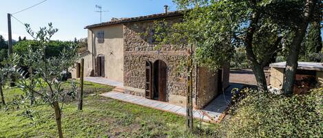 Un edificio de piedra con techo de tejas, acceso empedrado y un pequeño jardín con árboles y arbustos.