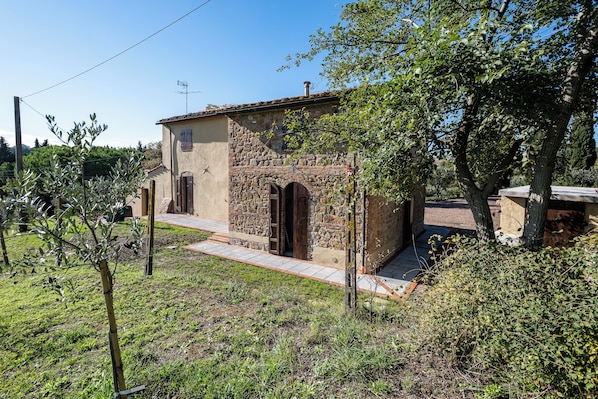 A stone building with a tiled roof, a paved entrance, and a small garden area with trees and shrubs.
