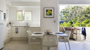 Eine moderne Küche mit weißen Schränken, ein Essbereich mit weißen Stühlen, Blick auf Grünflächen außerhalb und ein gerahmtes Bild an der Wand.