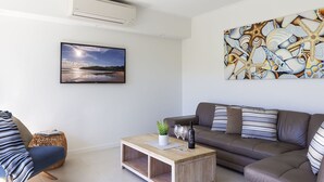 Ein modernes Wohnzimmer mit einer Couch, einem Couchtisch mit Pflanze und Weingläsern, einem gerahmten Landschaftsbild und einer dekorativen Wandgestaltung.