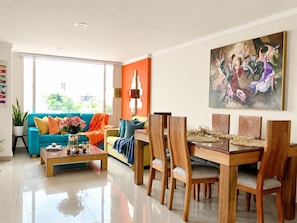 A modern living room with a teal sofa, a dining table with six chairs, and a large painting on the wall.