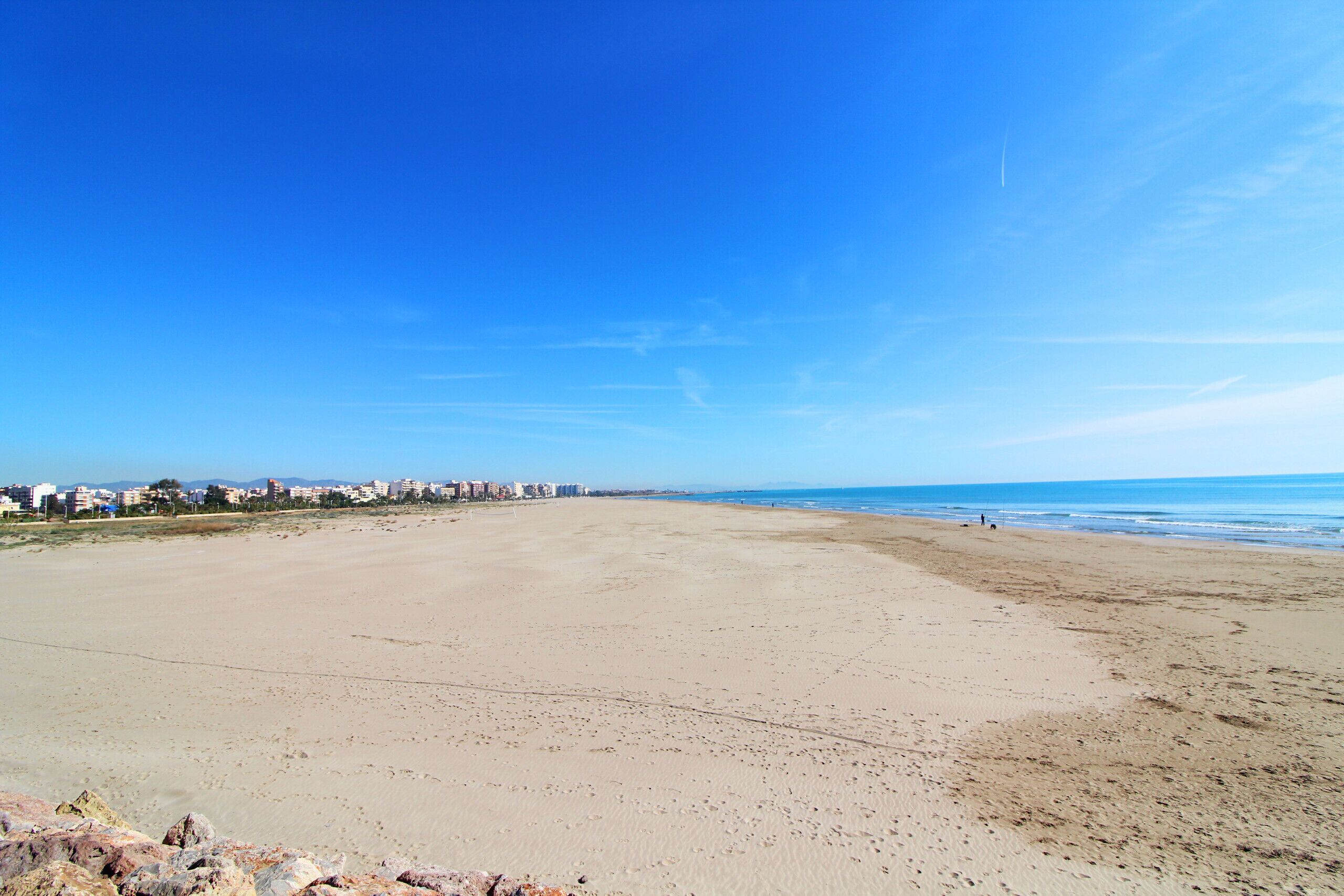 Una playa extensa con cielo despejado, una costa lejana con edificios y un primer plano rocoso.