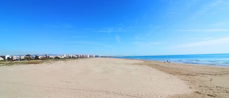 Una playa extensa con cielo despejado, una costa lejana con edificios y un primer plano rocoso.