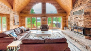L’intérieur d’une cabane en rondins est agrémenté d’une cheminée en pierre, de canapés en cuir et de grandes fenêtres qui offrent une vue sur l’extérieur.