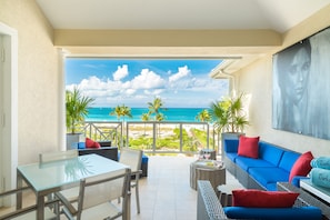 Relax in this serene oceanfront seating area with a glass-top table, sofa, and wicker chair, surrounded by lush greenery and a beautiful ocean view.