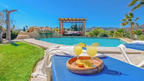 A poolside area with lounge chairs, a wooden serving tray with drinks, and a pergola in the background.