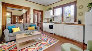 Una sala de estar con sofá, mesa de centro y alfombra colorida. Hay una zona de cocina con armarios, estufa y refrigerador.