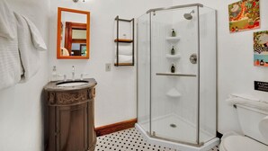 Un baño con ducha envuelta en vidrio, tocador de madera con lavamanos, inodoro blanco y espejo con marco de madera.