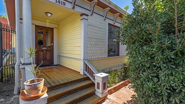 Una casa amarilla con el número 1419, una puerta principal de madera y una planta en maceta en los escalones.
