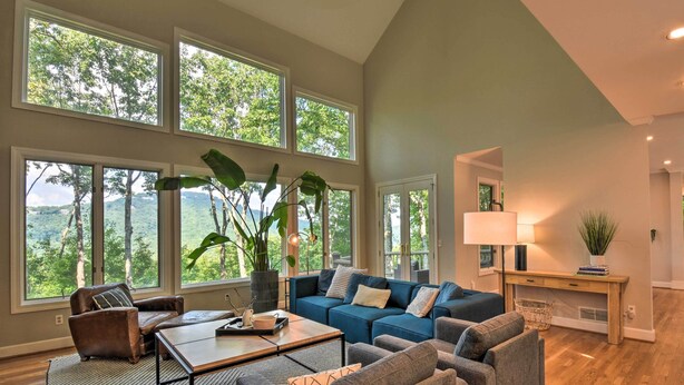 A spacious living room with large windows, a blue sectional sofa, a brown leather armchair, and a coffee table. - Luxe Big Canoe Resort Home w/ 3 Decks & Mtn Views! (Jasper)