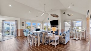 A spacious living area with a wooden floor, a ceiling fan, a dining table, and a sofa. - 1755 - Don't Worry Beach Happy - Walk to Beach | Pool, Hot Tub & Tiki Bar (Corolla)