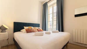 Una cama bien tendida con sábanas blancas y almohadas decorativas, una lámpara de noche y una ventana con cortinas.
