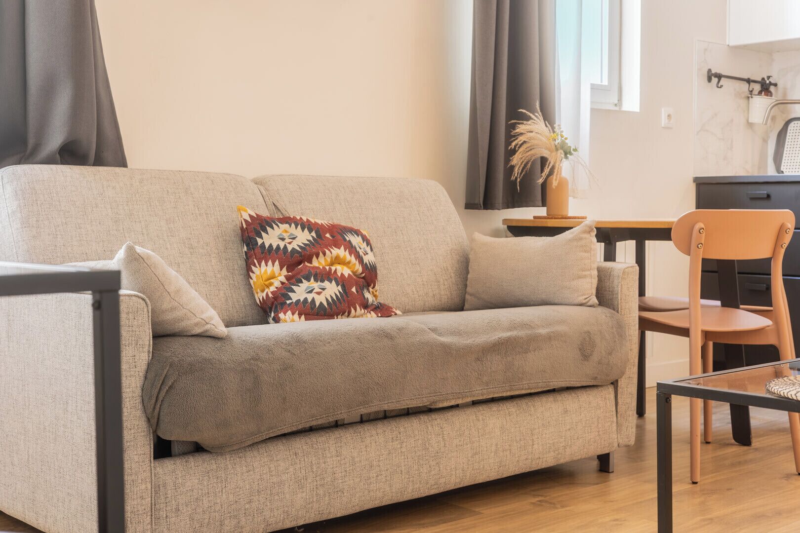 Un canapé aux couleurs neutres agrémenté d’un coussin à motifs, une chaise en bois et une petite table sur laquelle est posé un vase contenant des plantes séchées.