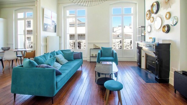 Una sala de estar con un sofá color turquesa, una mesita redonda pequeña y un sillón color turquesa. Hay una chimenea con una repisa negra y un espejo encima.