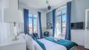 Habitación de hotel con cama, escritorio, silla, televisor y balcón con cortinas azules.