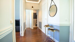 Un pasillo con suelo de madera, paredes blancas y una puerta que da a un baño. Hay dos pequeñas mesas redondas con marcos dorados y un espejo redondo en la pared.