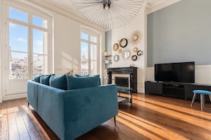 Un salon avec un canapé bleu, une cheminée, un téléviseur et un balcon doté de volets blancs.