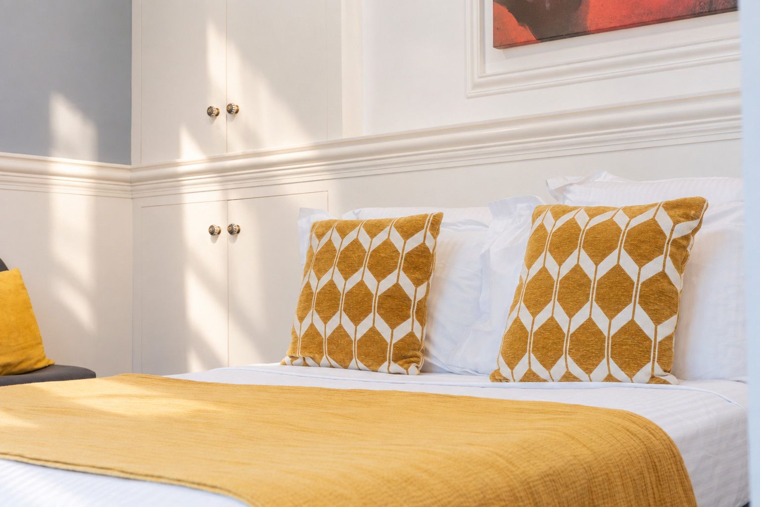 Ein Hotelzimmer mit einem Bett, zwei Kissen mit geometrischem Muster, einer gelben Decke und einer weißen Wand mit einem Gemälde.