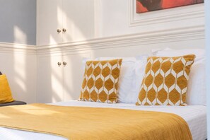 A hotel room with a bed, two pillows with a geometric pattern, a yellow throw blanket, and a white wall with a painting.
