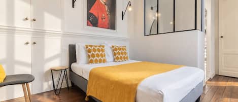 A bedroom with a bed, a chair, a small table, a painting, and a mirror.