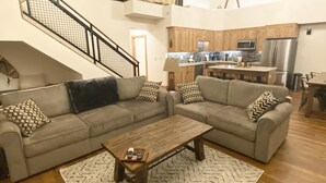 Un salón amplio con un sofá seccional gris, una mesa de centro de madera y un tapiz estampado. Se aprecia una cocina al fondo con armarios de madera y electrodomésticos modernos.
