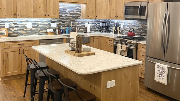 Una cocina moderna con armarios de madera, una isla central con taburetes de bar, electrodomésticos de acero inoxidable y un salpicadero de azulejos.