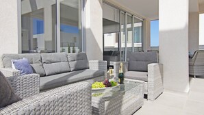Un área de estar moderna al aire libre con mobiliario de mimbre, una mesa de vidrio con frutas y bebidas, y vistas de la ciudad a través de amplios ventanales.