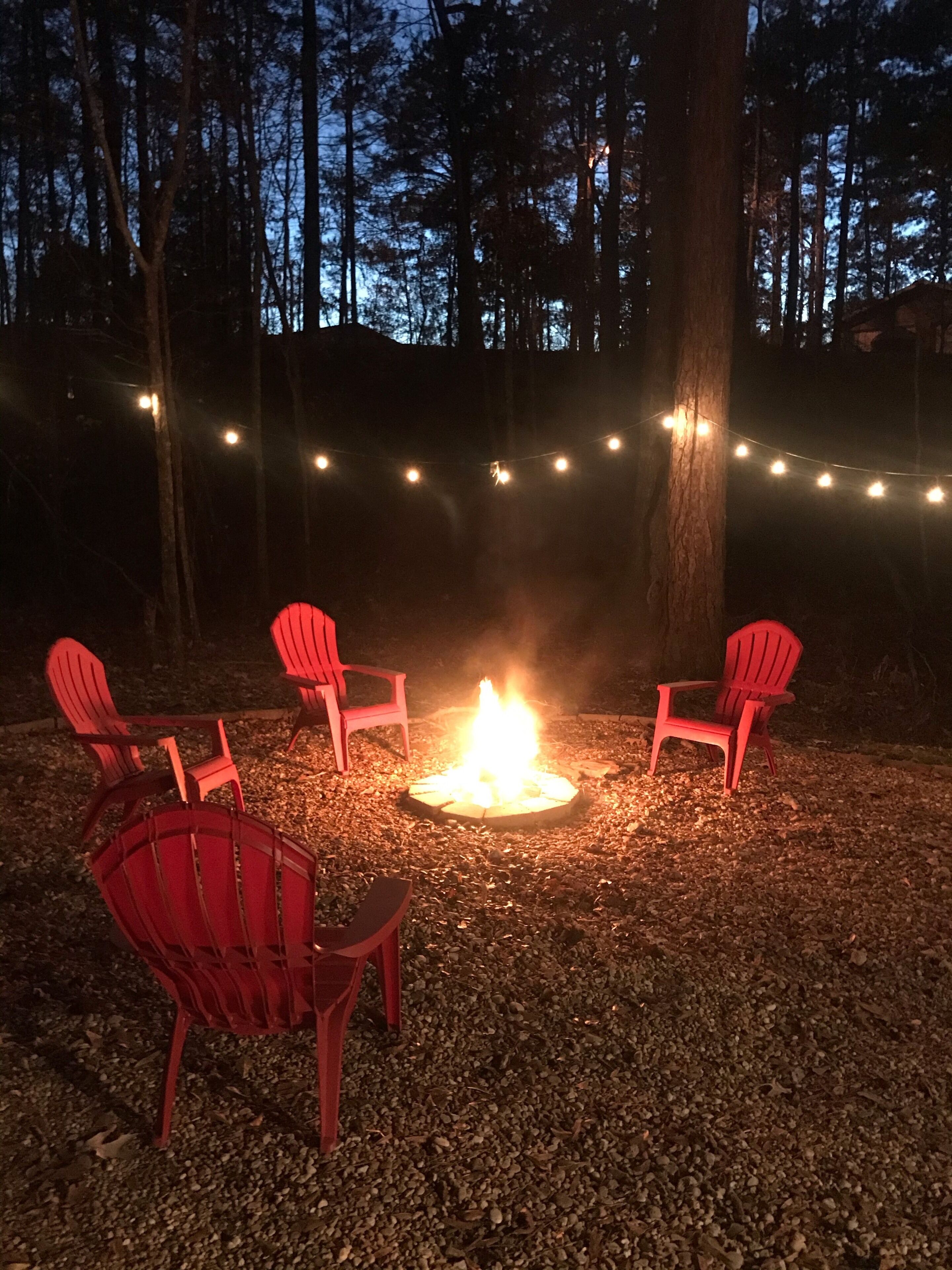 A campfire surrounded by red chairs in a forested area with string lights.