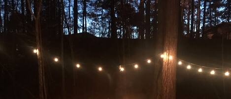 A campfire surrounded by red chairs in a forested area with string lights.