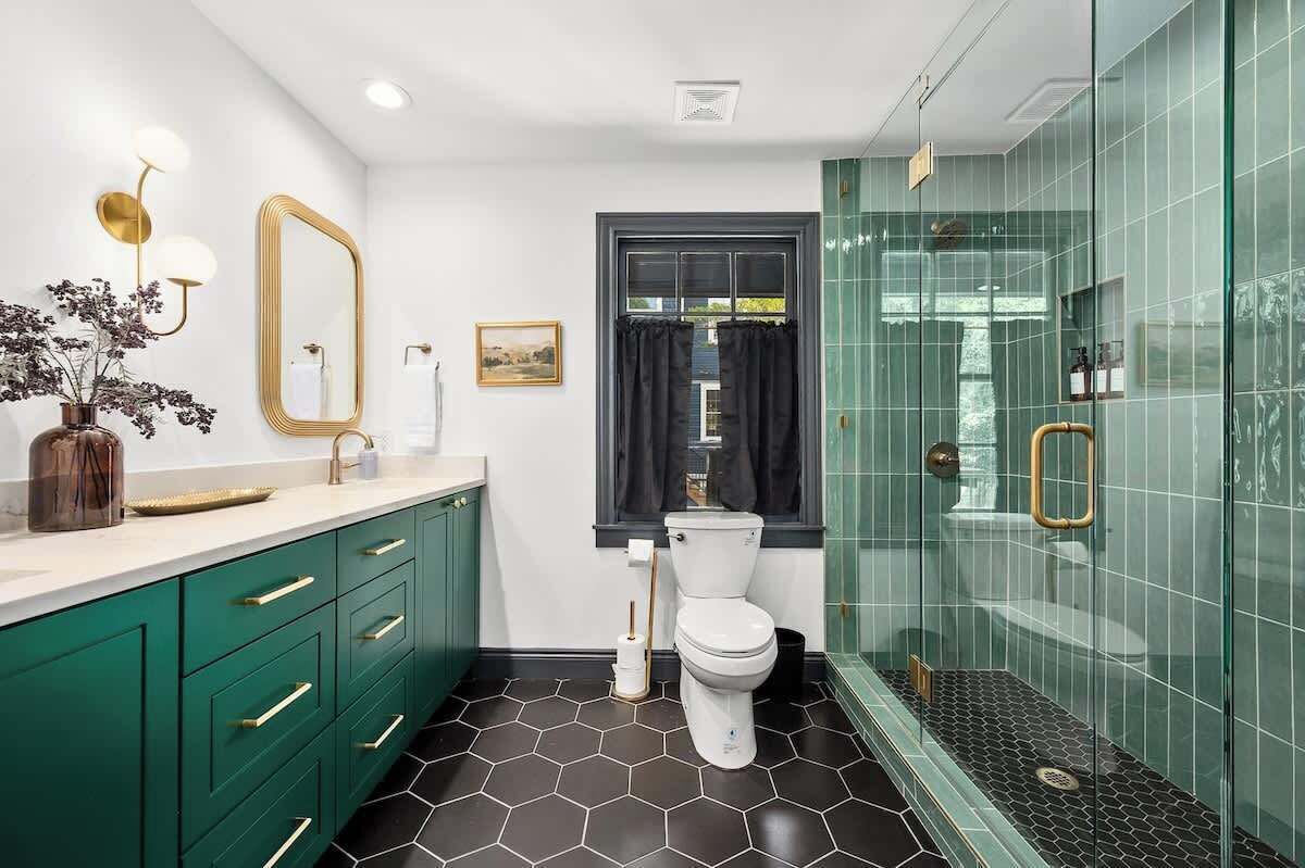 A modern bathroom with green cabinets, a white toilet, a glass shower enclosure, and a hexagonal floor pattern.