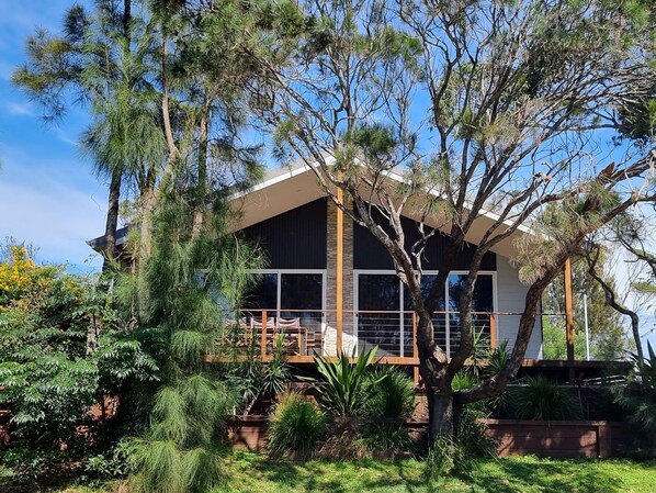 A modern house with a large deck, surrounded by lush greenery and tall trees. - Stylish modern open‑plan beach house offering a warm, welcoming, home‑like vibe. (Manyana)
