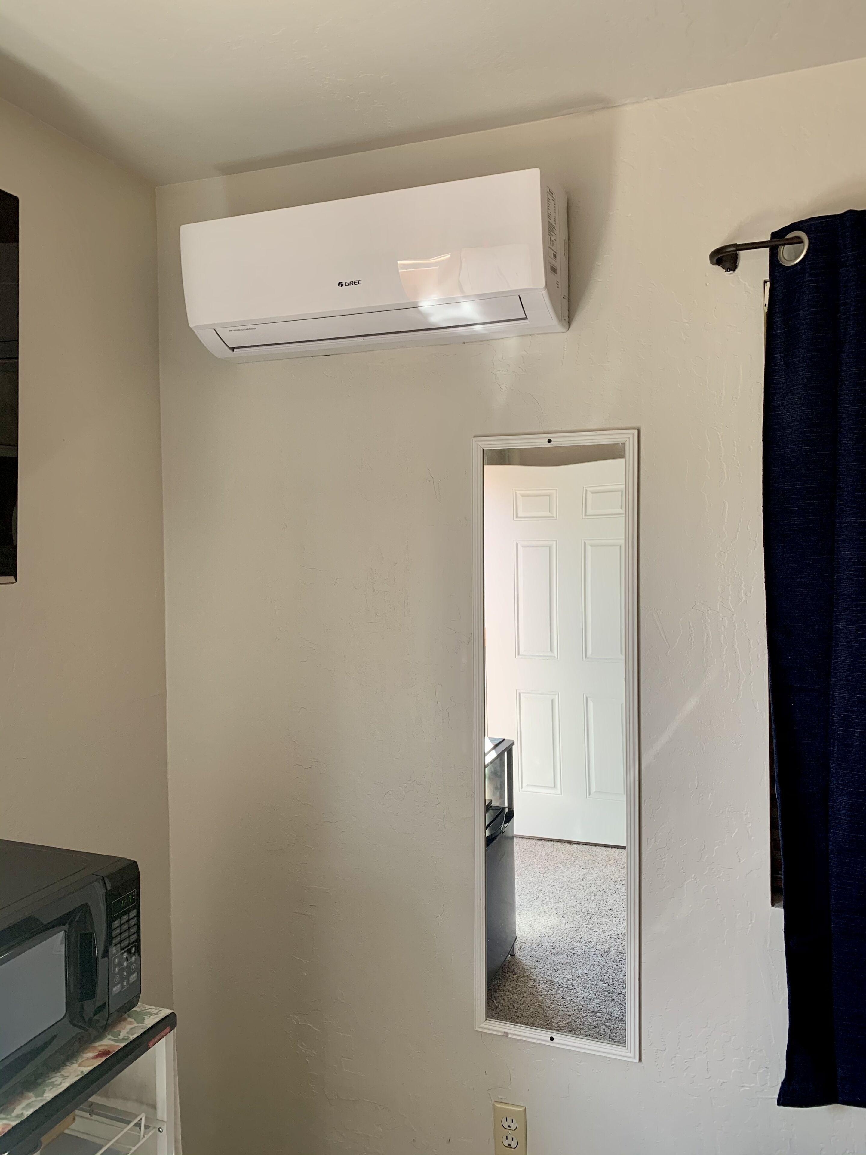 A room with a white air conditioner, a door, a dark curtain, and a microwave on a shelf.