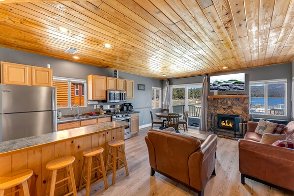 A modern kitchen with wooden cabinets, a central island with stools, a stone fireplace, and a view of the ocean through large windows.