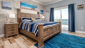 Un dormitorio con una cama de madera, una mesita de noche, una alfombra azul y vistas al océano a través de un ventanal grande.