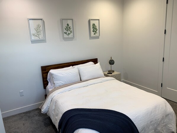 A neatly made bed with white bedding and a dark blue throw blanket, a bedside lamp, and three framed botanical prints on the wall.