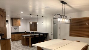 A modern kitchen with wooden cabinets, a central island, and a dining area with a white table and brown chairs. - Zion Canyon Retreat - Private Pool & Pickleball Court, Spacious, Clean & FUN!!! (La Verkin)