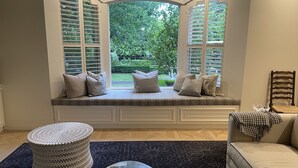 Ein Erker mit integrierten Fensterläden, eine gepolsterte Sitzbank mit Kissen, ein runder Hocker, ein Glastisch und ein Sofa mit Überwurfdecke.