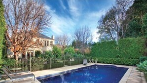 Ein Schwimmbecken mit gewölbter Kante, umgeben von einer Naturstein-Terrasse, einem Metallzaun und einer Hecke.