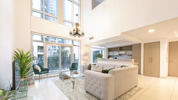 Ein geräumiger Wohnbereich mit einem weißen Sofa, einem Glastisch und einem grünen Sessel. Ein großes Fenster lässt Tageslicht herein und bietet einen Ausblick nach draußen.