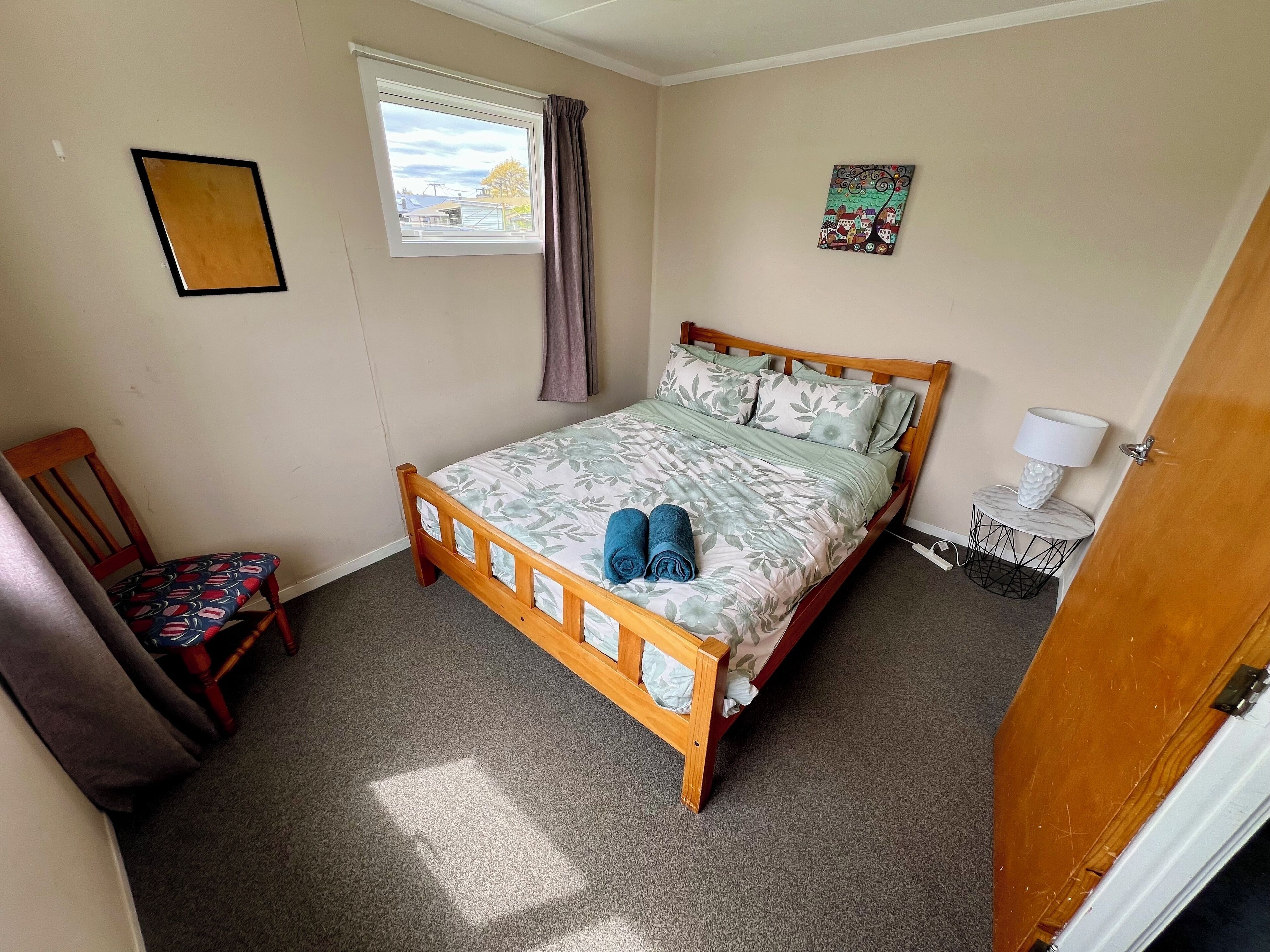 Une chambre à coucher avec un lit en bois, une chaise, une petite table avec une lampe et une fenêtre avec des rideaux.